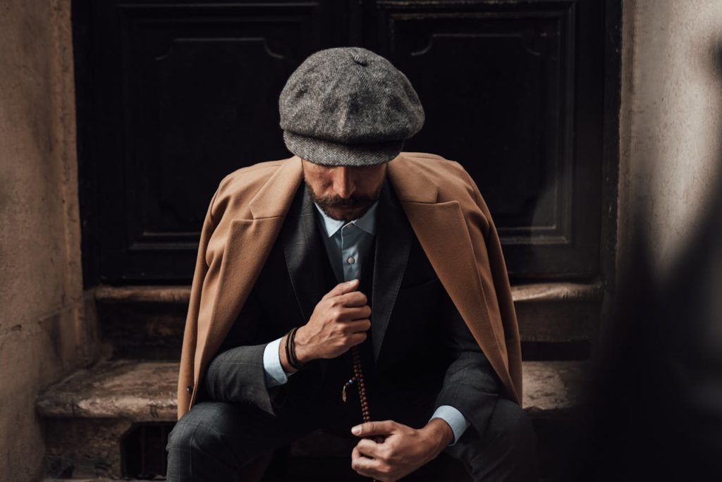 Pensive stylish male in posh suit coat and hat sitting with beads on shabby doorway and looking down in thoughts